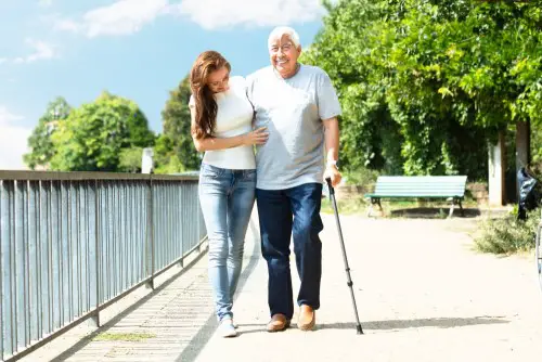 Young Woman Assisting Her Happy Father While Walking Near Railing In Park